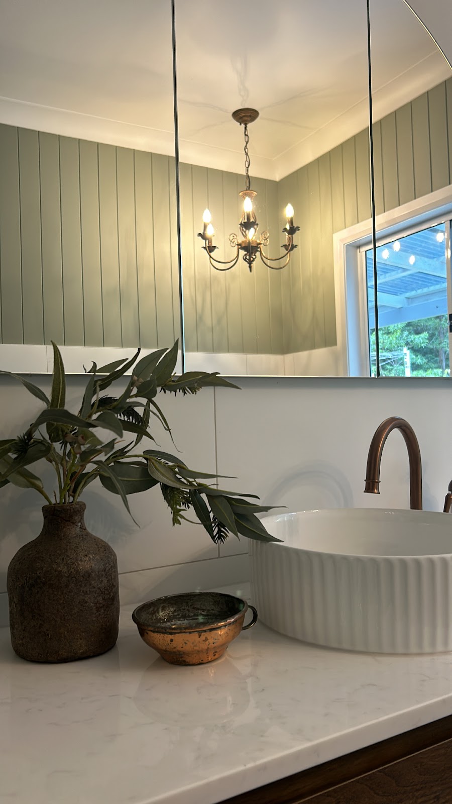 Fluted basin and copper tap on a white stone top, chandelier reflected in the mirror